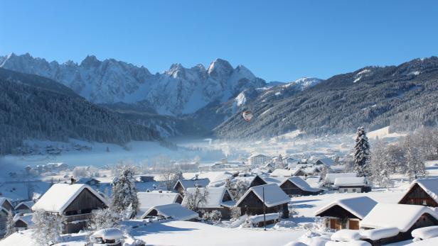 Winterliche Landschaft mit schneebedeckten Häusern und Bergen im Hintergrund. Ein Heißluftballon schwebt in der Ferne.