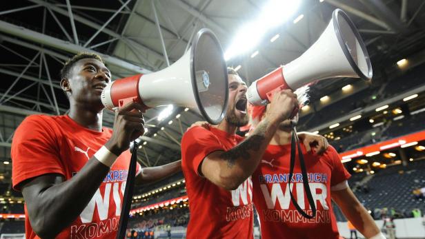 Drei Männer in roten T-Shirts jubeln mit Megaphonen in einem Stadion.