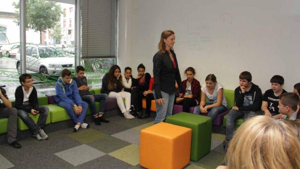 Eine Lehrerin steht vor einer Gruppe von Schülern, die auf bunten Sitzgelegenheiten sitzen.