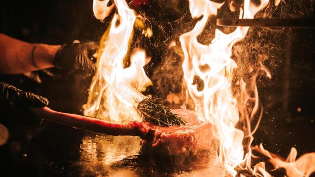 Ein Koch flambiert ein großes Steak mit Rosmarin auf einer heißen Platte.