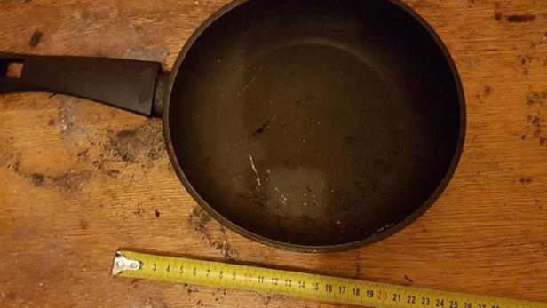 Eine Bratpfanne liegt neben einem gelben Maßband auf einem Holztisch.