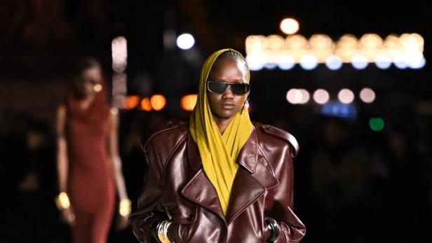 Eine Frau mit Sonnenbrille und Kopftuch trägt eine Lederjacke über einem gelben Kleid auf dem Laufsteg.