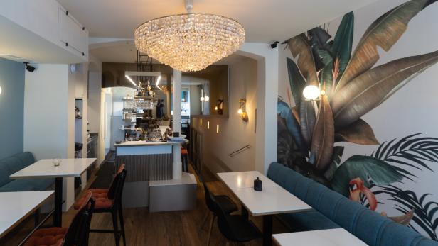 Innenansicht eines Restaurants mit Tischen, Stühlen, einer Bar und einer großen Kristalllampe.