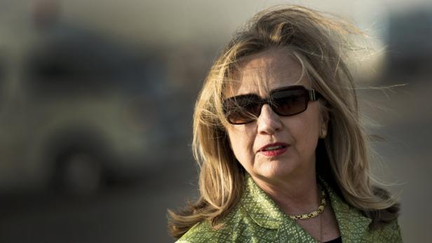 Hillary Clinton mit Sonnenbrille und grünem Blazer vor einem unscharfen Hintergrund.