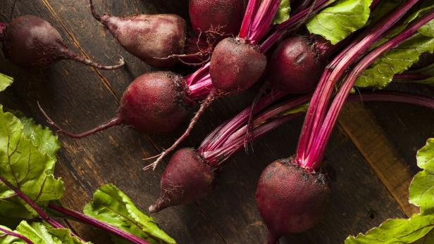 Eine Gruppe frischer, roter Rüben mit Blättern liegt auf einer dunklen Holzoberfläche.