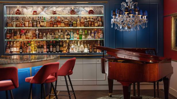 Eine Bar mit Spirituosenregal, roten Barhockern und einem Klavier unter einem Kronleuchter.