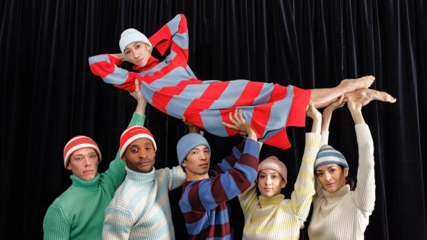 Eine Gruppe von Menschen in farbenfrohen Pullovern und Mützen trägt eine Person in einem rot-blau gestreiften Kleid.