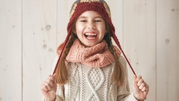 Ein lachendes Mädchen mit Zöpfen, Mütze und Schal vor hellem Hintergrund.