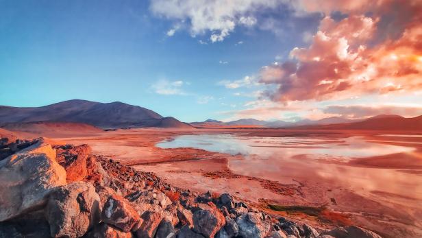 Eine rötliche Ebene mit einem See und Bergen unter einem bewölkten Himmel.