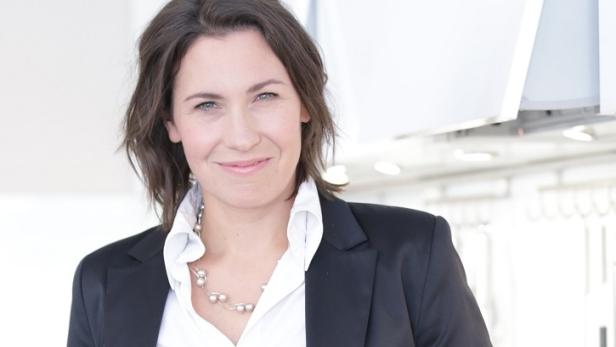 Eine Frau mit dunklen Haaren und einem schwarzen Blazer lächelt in die Kamera.