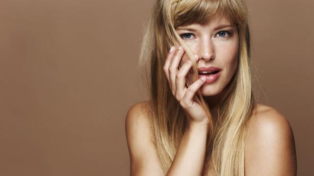 Eine blonde Frau mit blauen Augen berührt ihr Gesicht mit der Hand.