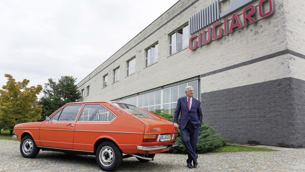 Giorgetto Giugiaro posiert neben einem orangefarbenen VW Passat vor dem Giugiaro-Designstudio.