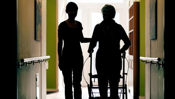 Eine junge Frau hilft einer älteren Dame mit einem Rollator in einem Flur.