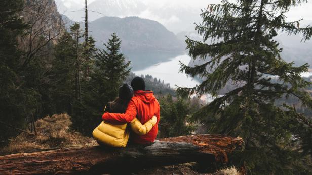 Ein Paar sitzt auf einem Baumstamm und blickt auf einen See in den Bergen.