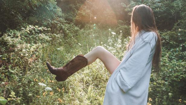 Eine Frau mit Morgenmantel und Cowboystiefeln steht in der Natur.