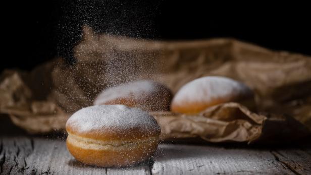 Drei Krapfen liegen auf einem Holztisch, während Puderzucker darüber gestreut wird.