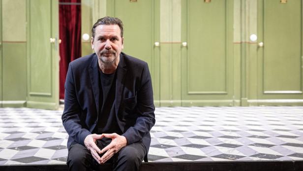 Ein Mann in einem dunklen Anzug sitzt vor grünen Türen und einem Schachbrettmusterboden.