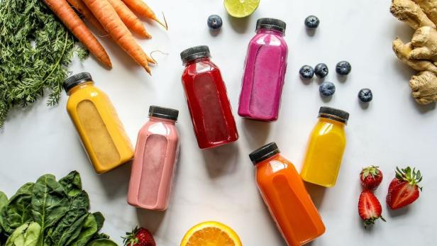 Mehrere bunte Saftflaschen sind mit Zutaten wie Karotten und Erdbeeren auf einer weißen Oberfläche arrangiert.