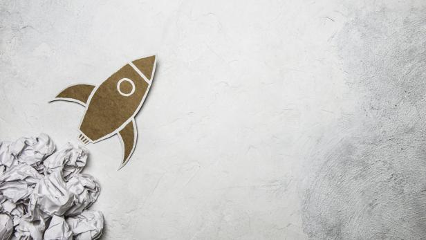 Eine Rakete aus Pappe hebt von zerknülltem Papier auf hellem Untergrund ab.