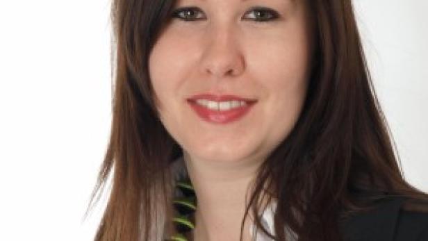 Eine lächelnde Frau mit braunen Haaren, Bluse und Blazer vor einem weißen Hintergrund.