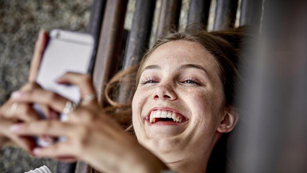 Laughing young woman lying on a bench using cell phone - Stock-Fotografie