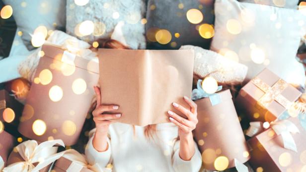 Beautiful woman is reading a book, laying between gift boxes, enjoying Christmas sale offers - Stock-Fotografie