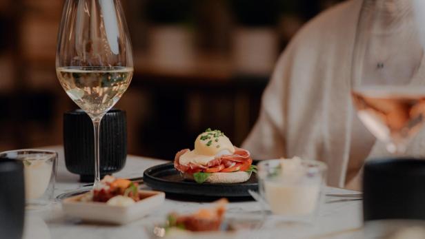 Ein gedeckter Tisch mit Speisen, darunter ein Glas Weißwein und Eggs Benedict.