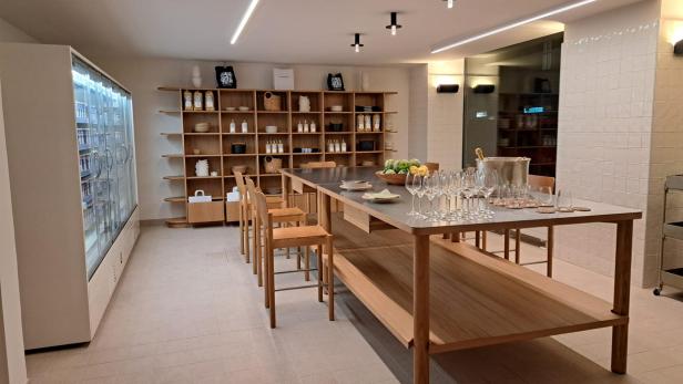 Ein langer Holztisch mit Stühlen in einem hellen Raum, im Hintergrund Regale und ein Kühlschrank.