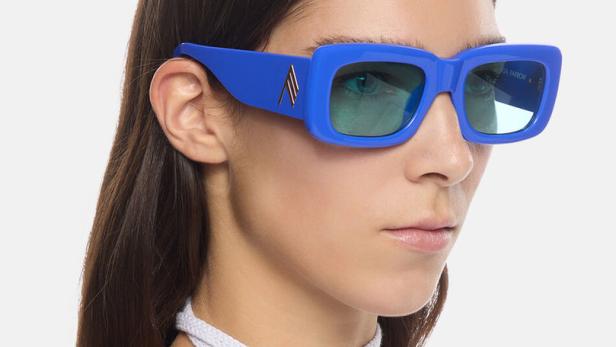 Eine Frau mit blauen, rechteckigen Sonnenbrillen und einem weißen Oberteil mit Schnürung.