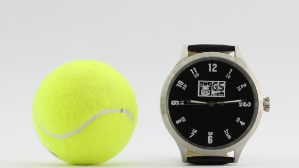 Ein Tennisball liegt neben einer Armbanduhr mit dem GS-Logo.