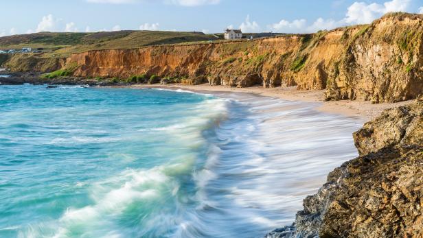Türkisfarbenes Wasser umspült einen Sandstrand mit Klippen im Hintergrund.