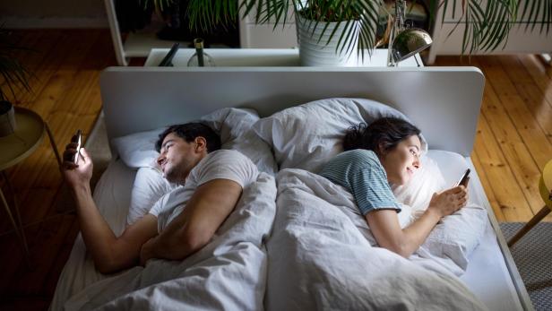 Ein Mann und eine Frau liegen Rücken an Rücken im Bett und benutzen ihre Mobiltelefone. Das Paar ignoriert sich und nutzt seine Smartphones.