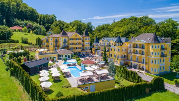 Ein gelbes Hotelresort mit Pool und gepflegten Gärten vor einem bewaldeten Hügel.