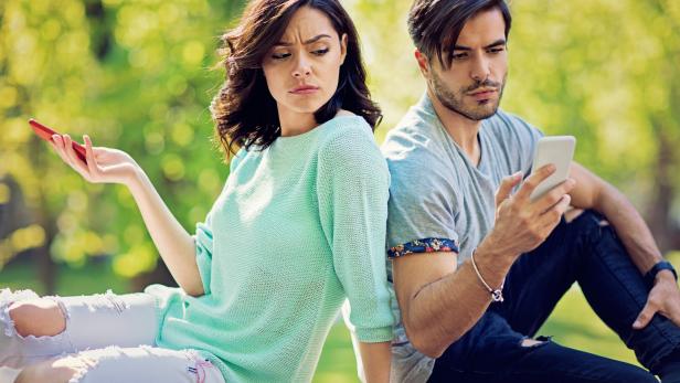 Ein Paar sitzt Rücken an Rücken im Park und schaut auf ihre Handys.