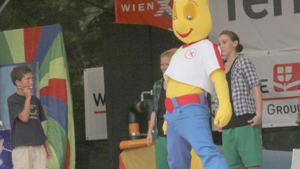 Ein gelbes Maskottchen steht mit Kindern auf einer Bühne.