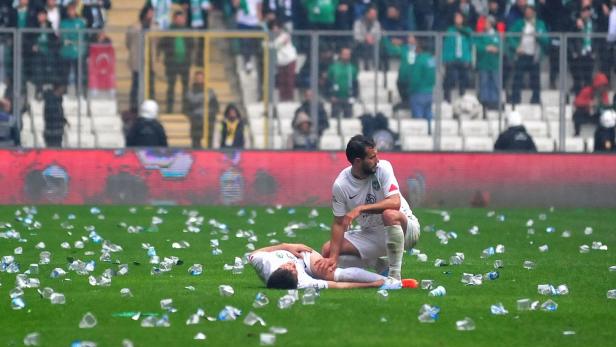 Ein Fußballspieler kniet neben einem verletzten Spieler auf einem mit Flaschen übersäten Feld.
