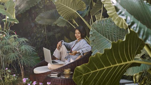 Eine asiatische Frau arbeitet von zu Hause aus, sitzt in Gartenmöbeln, umgeben von tropischen Pflanzen, und benutzt einen Laptop und ein Mobiltelefon.