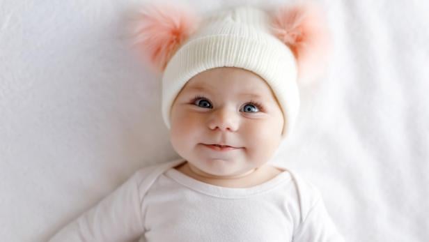 Süßes entzückendes Babykind mit warmweißem und rosa Hut mit süßen Bobbles. Glückliches kleines Mädchen auf weißem Hintergrund und Blick in die Kamera. Nahaufnahme für Weihnachtsurlaub und Familienkonzept.