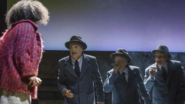 Eine Frau steht vor drei Männern mit Hüten und Anzügen auf einer Theaterbühne.