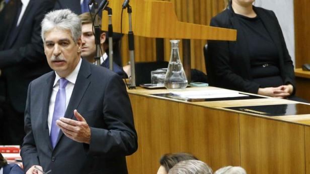 Ein Mann mit grauem Haar und Schnurrbart spricht in einem Parlament.
