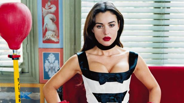 Monica Bellucci posiert mit Boxhandschuhen auf einem roten Sofa.