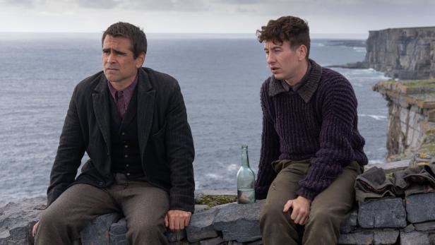 Colin Farrell und Barry Keoghan sitzen mit einer Flasche auf einer Steinmauer vor einer Küstenlandschaft.