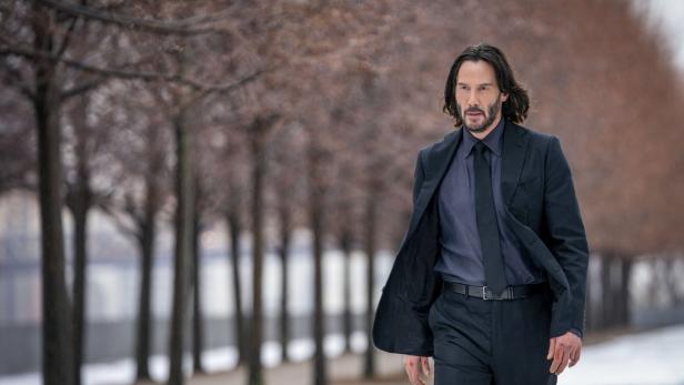 Keanu Reeves in einem dunklen Anzug geht auf einem verschneiten Weg entlang einer Baumreihe.