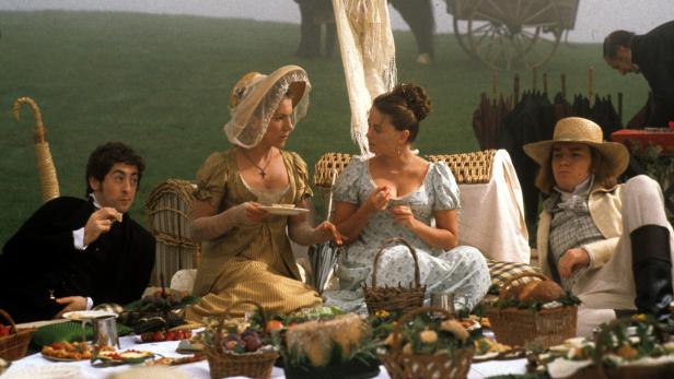 Eine Picknickgesellschaft in Kostümen des frühen 19. Jahrhunderts sitzt auf einer Wiese.