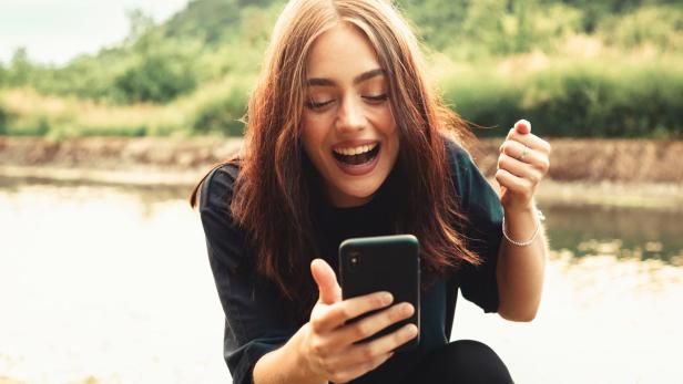 Eine junge Frau jubelt, während sie auf ihr Smartphone schaut. (Symbolbild).