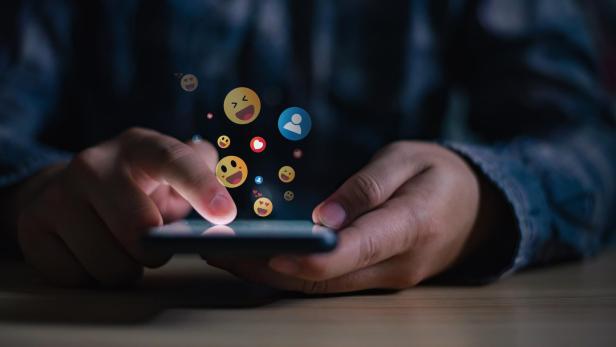 Ein junger Mann tippt auf sein Smartphone, wobei verschiedene positive Emojis auf dem Bildschirm erscheinen.