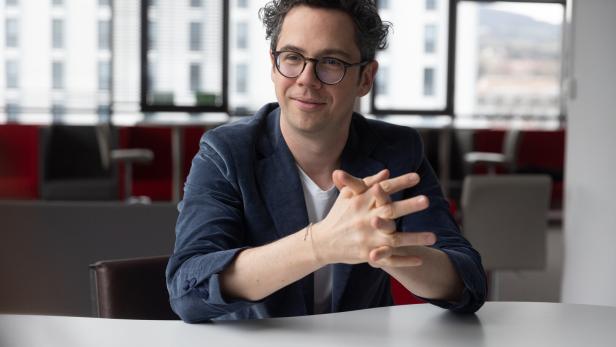 Ein Mann mit Brille und lockigen Haaren sitzt lächelnd an einem Tisch in einem modernen Büro.