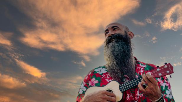 Rasierter Mann mit langem Bart spielt Ukulele vor einem dramatischen Himmel.