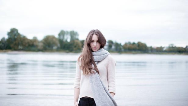 Eine junge Frau mit langem Haar und einem Schal steht vor einem See.
