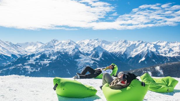 Zwei Personen entspannen auf grünen Sitzsäcken im Schnee vor einer Bergkulisse.